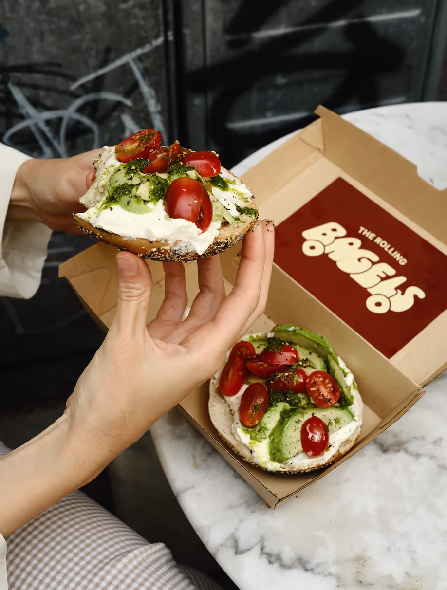 Hands holding a Rolling Bagels bagel with avocado, tomato and pesto over an open box
