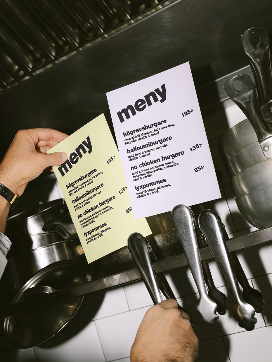 Hands holding yellow and lilac HK's Lila menus over kitchen pots
