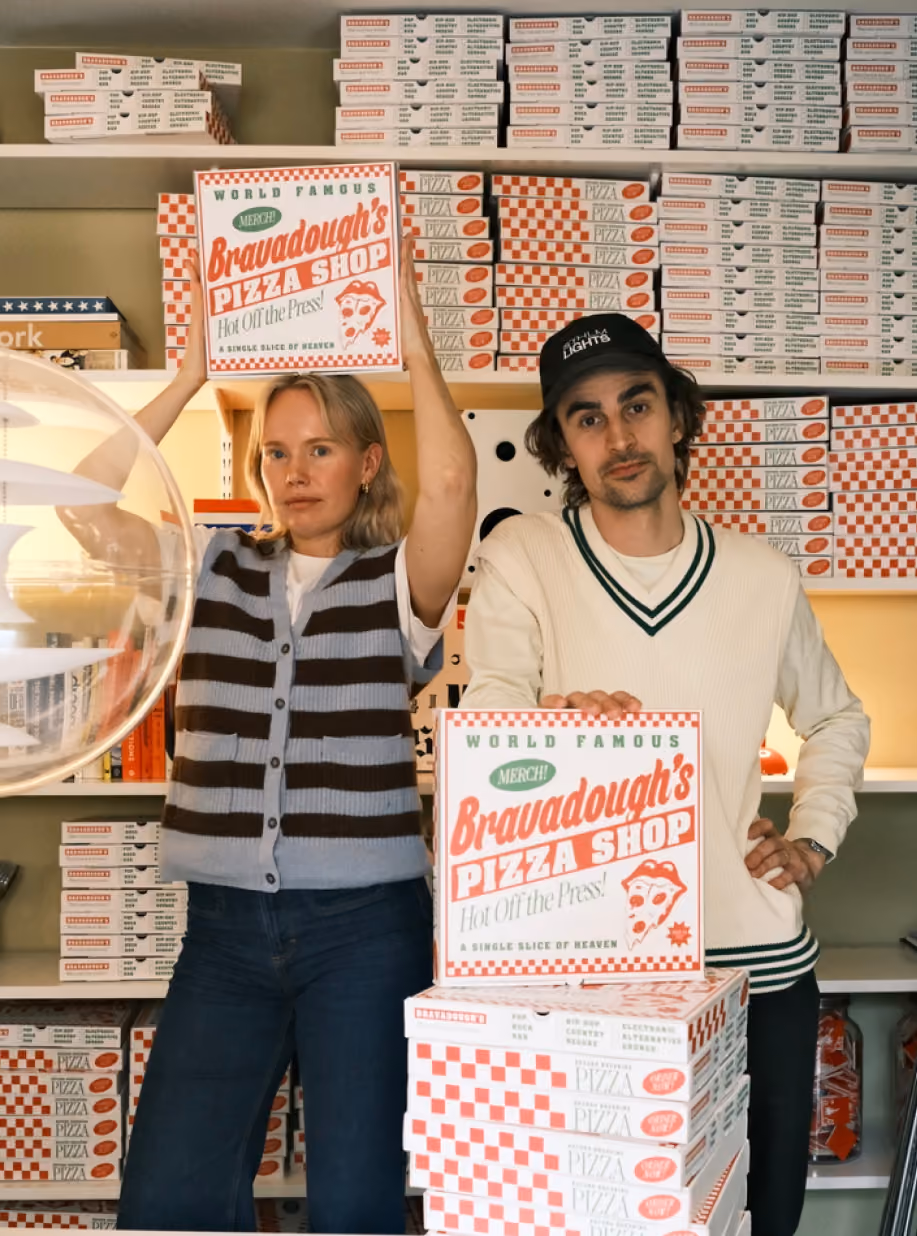 Two hosts holding Bravadough's Pizza Shop boxes in the slice-shop set